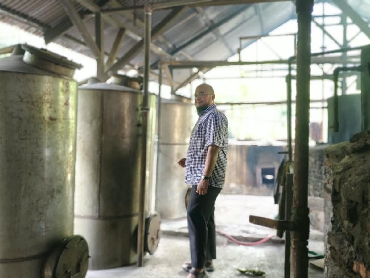 Hasan standing beside tall steel vessels inside a distillery shed